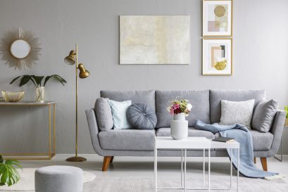 Gold mirror above shelf with plant in grey living room interior grey sofa and flowers on table